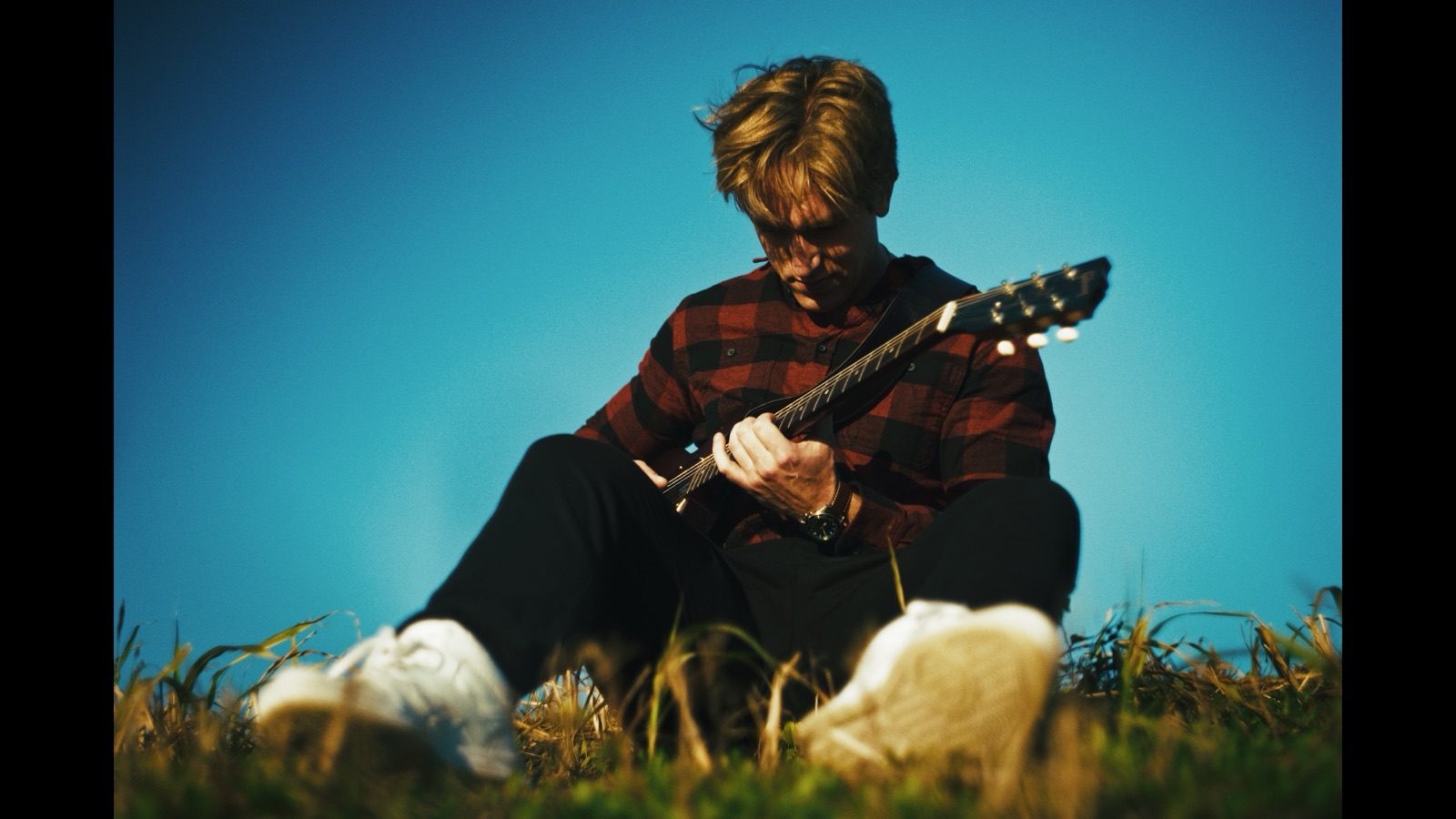 Nick sitting in a field playing electric guitar under a bright blue sky